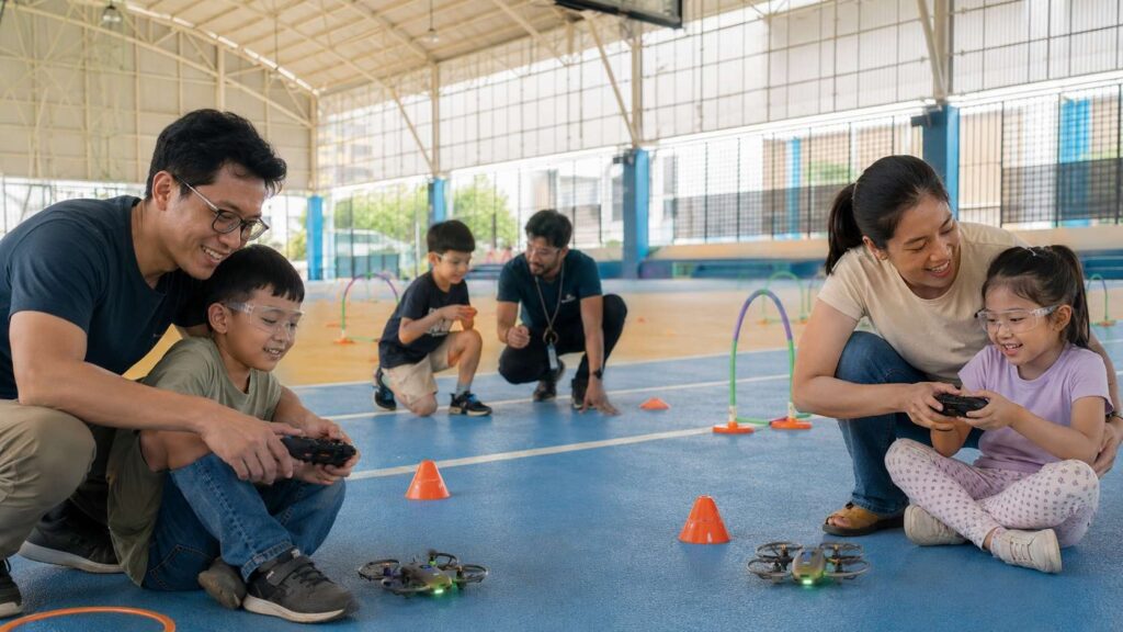 Parents and children learning to fly small drones together during a hands-on STEM workshop in an indoor sports hall with safety goggles, controllers, cones, and obstacle gates.